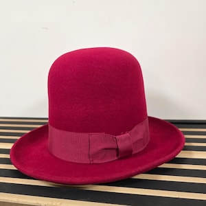 Può includere: Un cappello bombetta rosso scuro con un nastro e un fiocco abbinati. Il cappello ha una corona arrotondata e un'ampia tesa. Il cappello è realizzato in un materiale simile al feltro e poggia su una superficie a strisce bianche e nere.