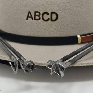 Puede incluir: Un sombrero de fieltro beige con las letras ABCD marcadas en la parte frontal, con una cinta de cuero negra y marr&oacute;n. Dos hierros de marcar de metal con formas de letras distintas se sit&uacute;an en primer plano, apoyados en el ala del sombrero.