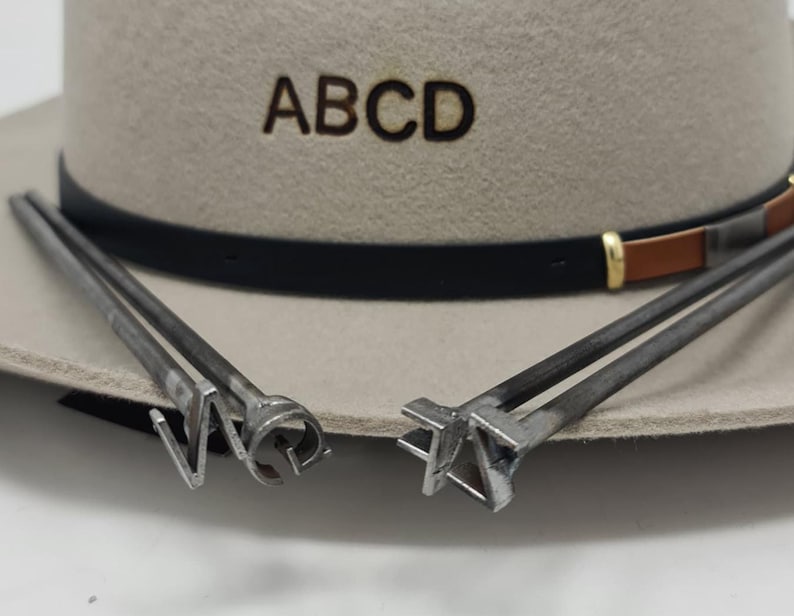 Puede incluir: Un sombrero de fieltro beige con las letras ABCD marcadas en la parte frontal, con una cinta de cuero negra y marr&oacute;n. Dos hierros de marcar de metal con formas de letras distintas se sit&uacute;an en primer plano, apoyados en el ala del sombrero.