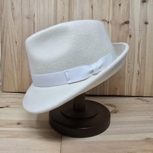 May include: A white fedora hat with a white ribbon band. The hat is sitting on a wooden hat stand.