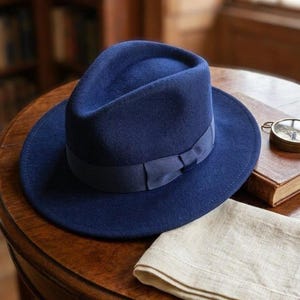 Könnte beinhalten: Ein tief dunkelblauer Filz-Fedora-Hut mit einem passenden Ripsband und einer kleinen Schleife. Der Hut hat eine strukturierte Krone mit einer Mittelkerbe und eine breite, flache Krempe, was eine klassische Silhouette und Textur ergibt.