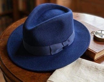 Op de afbeelding: Een diep marineblauwe vilten fedora-hoed met een bijpassende grosgrain lintband met een klein strikdetail. De hoed heeft een gestructureerde kroon met een middenvouw en een brede, platte rand, wat zorgt voor een klassiek silhouet.