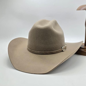 Puede incluir: Un sombrero de vaquero beige con ala ancha y copa redondeada. El sombrero presenta una banda decorativa con una hebilla plateada. El sombrero es de material similar al fieltro y se exhibe en un soporte de madera.