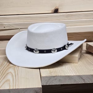 May include: A white felt cowboy hat with a black band and silver accents. The hat has a wide brim and a tall crown.