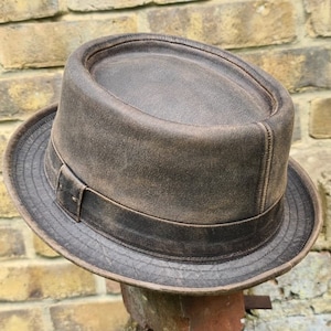 Könnte beinhalten: Ein brauner Lederhut im Fedora-Stil mit abgenutzter Oberfläche. Der Hut hat eine breite Krempe und eine flache Krone.