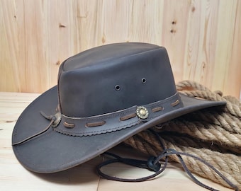 May include: A brown leather cowboy hat with a wide brim and a braided leather band. The hat has a decorative metal buckle and a chin strap.