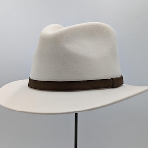 Puede incluir: Un sombrero fedora blanco con una banda de cuero marrón.