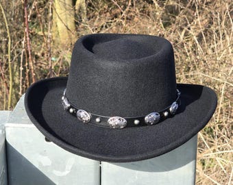 Może przedstawiać: Czarny filcowy kapelusz fedora z szerokim rondem i srebrnym pasem z konchami.