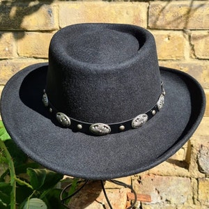 Könnte beinhalten: Ein schwarzer Filzhut im Fedora-Stil mit breiter Krempe und einem silbernen und schwarzen Band mit dekorativen Nieten.