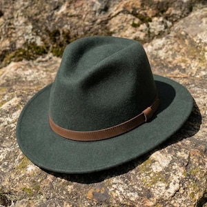 May include: A dark forest green fedora hat featuring a structured crown and a wide brim. The hat is accented with a smooth, thin brown leather band that encircles the base of the crown, providing a distinct contrast to the deep green felt material.