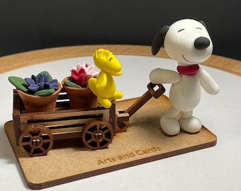 May include: A white dog figurine with a red collar pulls a wooden wagon with two potted plants and a yellow bird figurine. The wagon has brown wheels and the base is made of wood with the text "Arts and Cards" engraved on it.