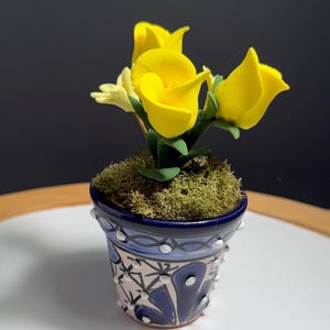 May include: A small, decorative potted plant featuring bright yellow calla lilies. The ceramic pot is painted with a blue and white floral design and accented with white dots. The plant sits on a bed of green moss.