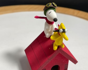 May include: A white dog wearing a green aviator hat and red scarf stands on the roof of a red wooden doghouse with a yellow bird wearing goggles.