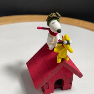 May include: A white dog wearing a green aviator hat and red scarf stands on the roof of a red wooden doghouse with a yellow bird wearing goggles.