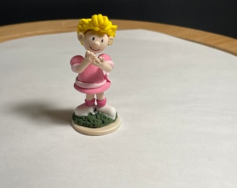 May include: A small figurine of a girl with yellow hair wearing a pink dress and white shoes. She is standing on a green base.
