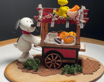 May include: A Snoopy and Woodstock figurine set. Snoopy, in a red collar, pushes a red and brown cart filled with pastries and a cup of coffee. Woodstock sits above the cart. The base is brown with green grass and the text "Arts and Cards".