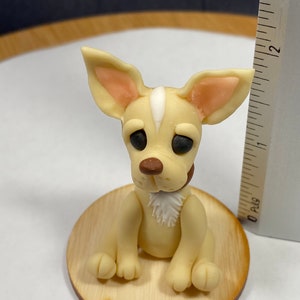 May include: A light tan chihuahua figurine with a white stripe on its head, brown nose, and black eyes. It sits on a small wooden base with the text "Arst and Cards". The figurine has a small brown collar and a white patch of fur on its chest.