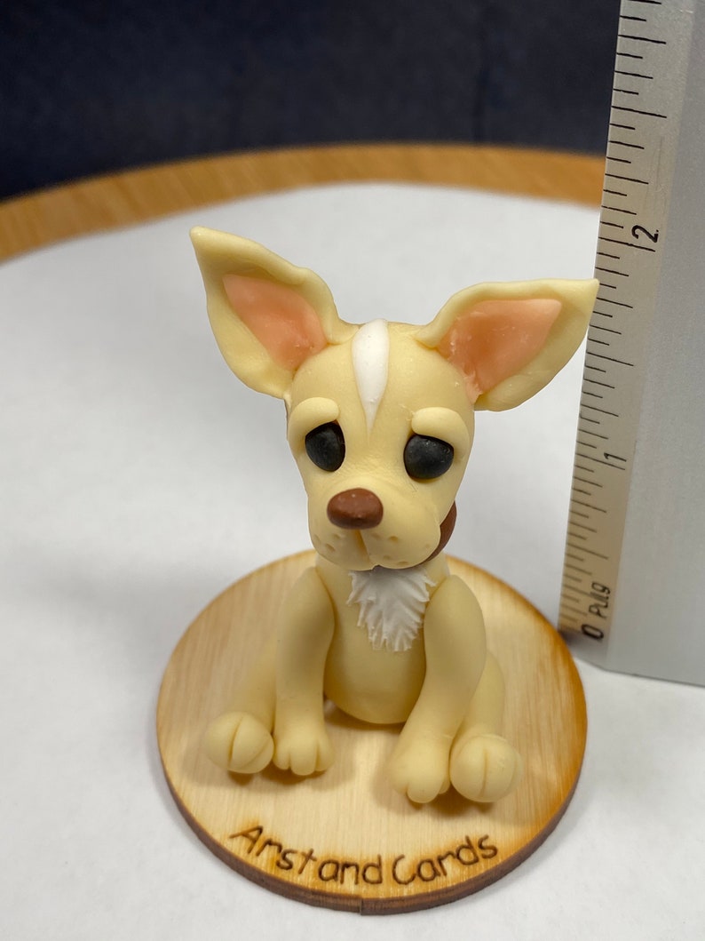 May include: A light tan chihuahua figurine with a white stripe on its head, brown nose, and black eyes. It sits on a small wooden base with the text "Arst and Cards". The figurine has a small brown collar and a white patch of fur on its chest.