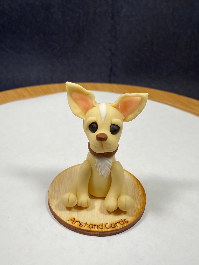 May include: A light tan Chihuahua figurine with a white stripe on its head, wearing a brown collar, and sitting on a wooden base. The base has the text "Arst and Cards" engraved on it. The dog has large, dark eyes and floppy ears.