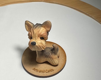 May include: A small ceramic figurine of a brown and black Yorkshire Terrier dog sitting on a round wooden base with the text "Arts and Cards" engraved on it.