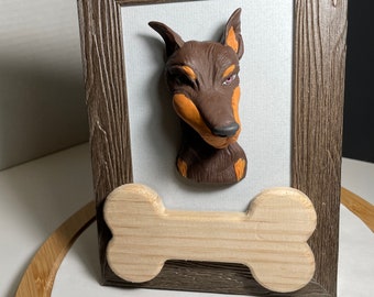 May include: A brown and black dog sculpture in a wooden frame with a wooden bone underneath. The dog has a serious expression and is looking to the right.