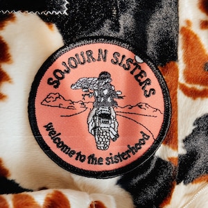 May include: A pink and black embroidered patch with the text "Sojourn Sisters" and "Welcome to the Sisterhood". The patch features a motorcycle with two riders and a mountain landscape.