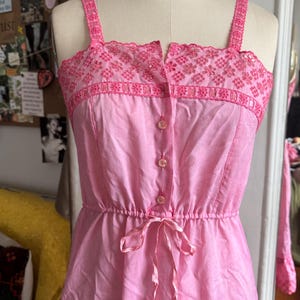 Puede incluir: Un vestido de verano rosa con detalles bordados en los tirantes y el corpiño. El vestido presenta una botonadura frontal, una cintura ceñida con una cinta y una falda fluida. El vestido se exhibe en un maniquí.