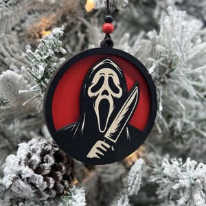 May include: A black and white wooden Christmas ornament featuring a ghost wearing a mask and holding a knife. The ghost is against a red background.
