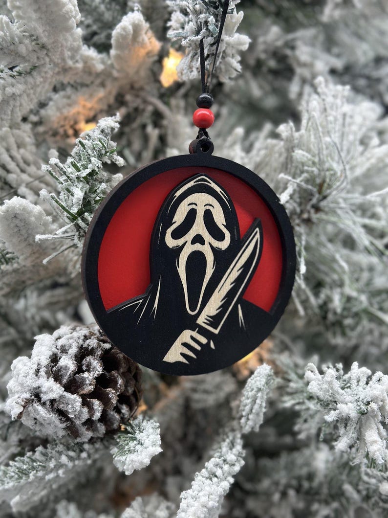 May include: A black and white wooden Christmas ornament featuring a ghost wearing a mask and holding a knife. The ghost is against a red background.