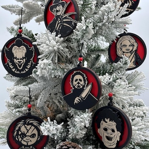 May include: Seven round, red and black, wooden ornaments featuring horror movie characters. The ornaments are hanging on a snowy artificial Christmas tree.