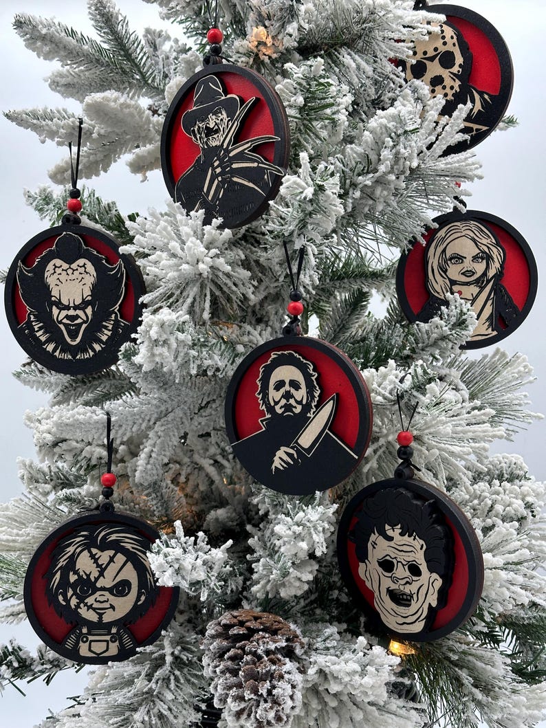 May include: Seven round, red and black, wooden ornaments featuring horror movie characters. The ornaments are hanging on a snowy artificial Christmas tree.