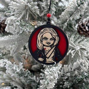 May include: A black and red wooden Christmas ornament featuring a character with blonde hair, a red dress, and a knife. The ornament is hanging on a snowy Christmas tree.