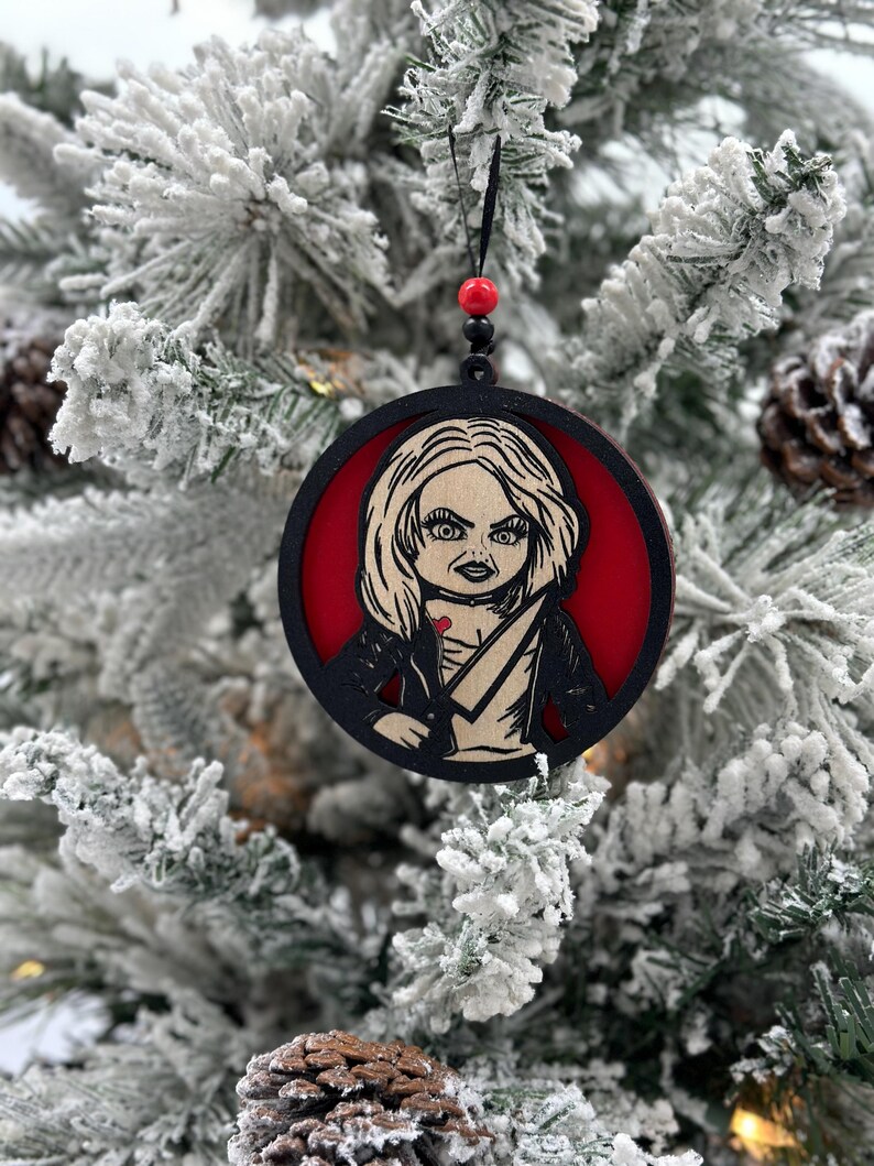 May include: A black and red wooden Christmas ornament featuring a character with blonde hair, a red dress, and a knife. The ornament is hanging on a snowy Christmas tree.