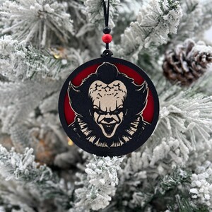 May include: A round wooden Christmas ornament with a black and white image of a scary clown with red eyes and a wide smile. The ornament is hanging from a black string with red beads.