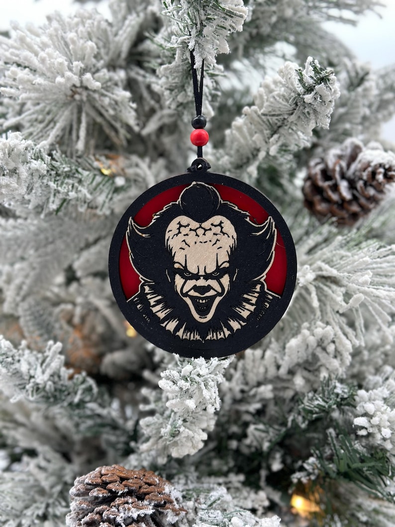 May include: A round wooden Christmas ornament with a black and white image of a scary clown with red eyes and a wide smile. The ornament is hanging from a black string with red beads.