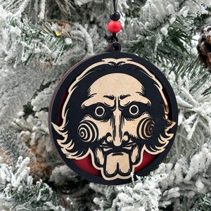 May include: A wooden Christmas ornament featuring a black and white illustration of a character from the horror movie "Saw". The ornament is round and has a red background.