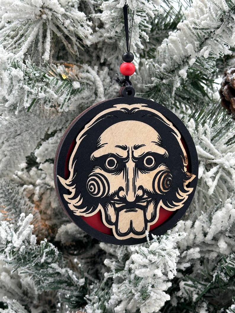 May include: A wooden Christmas ornament featuring a black and white illustration of a character from the horror movie "Saw". The ornament is round and has a red background.