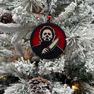 May include: A black and red round Christmas ornament featuring a silhouette of a horror movie character with a knife. The ornament is hanging on a snowy Christmas tree.