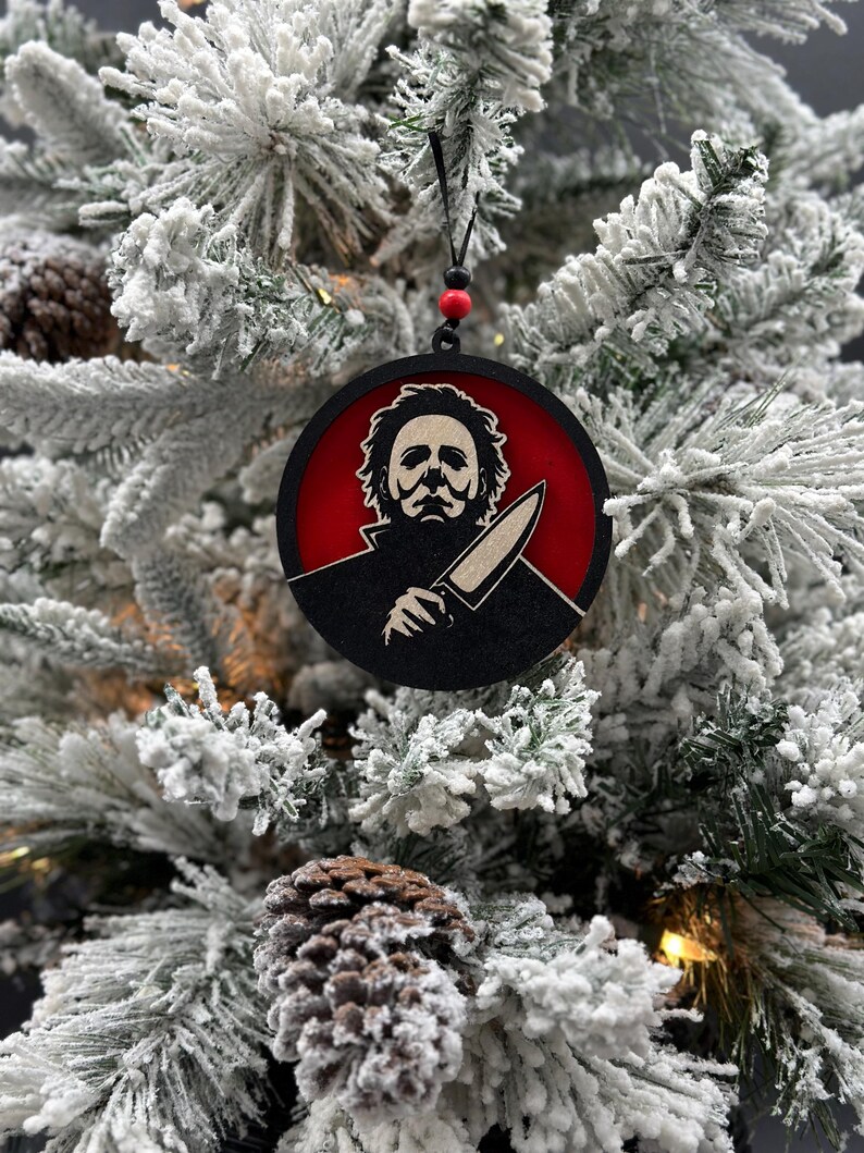 May include: A black and red round Christmas ornament featuring a silhouette of a horror movie character with a knife. The ornament is hanging on a snowy Christmas tree.