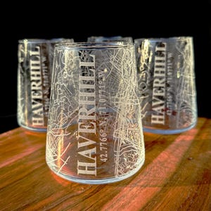 May include: A set of clear stemless glasses with a map design etched into the glass. The word "HAVERHILL" is vertically printed on each glass, along with the coordinates 42.7766° N, 71.0771° W. The glasses are displayed on a wooden surface.