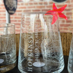 Puede incluir: Vasos de vidrio transparente con un mapa de Hopkinsville, KY grabado en la superficie. El texto "HOPKINSV" y las coordenadas 86.56°N 87.4886°W son visibles. Un marcador de bebida en forma de estrella de mar rosa brillante está adherido a un vaso.