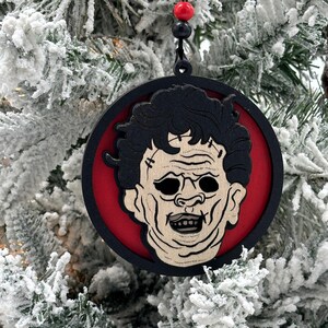 May include: A round wooden Christmas ornament featuring a black and white illustration of a horror movie character's face. The ornament is painted red and black and has a black string for hanging.