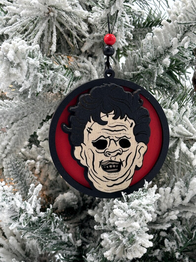 May include: A round wooden Christmas ornament featuring a black and white illustration of a horror movie character's face. The ornament is painted red and black and has a black string for hanging.