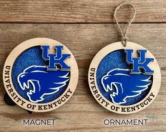 May include: A circular wooden magnet and a circular wooden ornament, each featuring a blue glitter background, a blue University of Kentucky wildcat logo, the text "UNIVERSITY OF KENTUCKY" in black, and a blue "UK" monogram. The ornament has a twine loop.