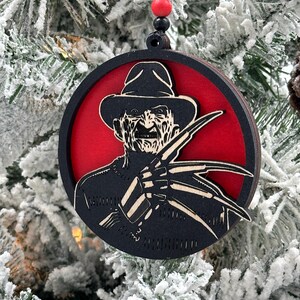 May include: A round, black and red wooden Christmas ornament featuring a silhouette of Freddy Krueger from the Nightmare on Elm Street movie franchise. The ornament has a black ribbon for hanging.