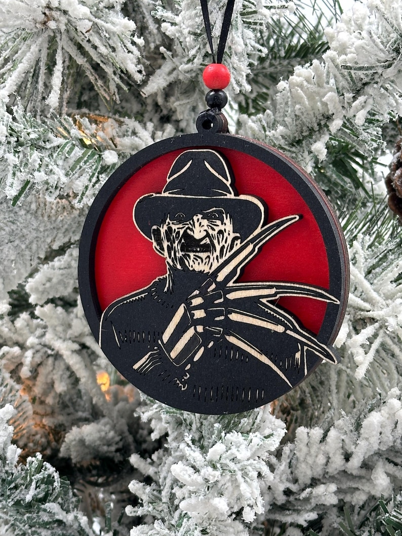 May include: A round, black and red wooden Christmas ornament featuring a silhouette of Freddy Krueger from the Nightmare on Elm Street movie franchise. The ornament has a black ribbon for hanging.