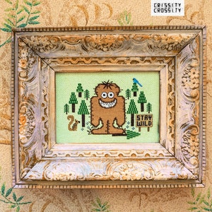 May include: Cross-stitch artwork in a distressed, ornate frame. The artwork features a brown monster, green trees, a blue bird, and the words "STAY WILD." The frame is cream-coloured with a distressed finish.