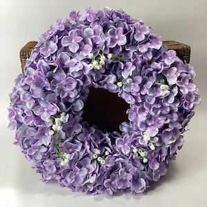 May include: A circular wreath of purple hydrangea flowers, with small white blossoms. The wreath is set against a brown woven background. A decorative piece for the home, bringing a touch of nature indoors.