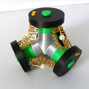 May include: A three-pronged mechanical object featuring a central silver Y-shaped core with bright green accents. Three black circular discs are connected to the core by gold-coloured metal springs, each adorned with a single white spherical bead.