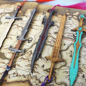 May include: Five sword-shaped bookmarks made of wood, each featuring distinct engraved details and a coloured tassel. The bookmarks are arranged on a printed map background. The designs include natural wood, grey, dark red, and bright turquoise finishes.